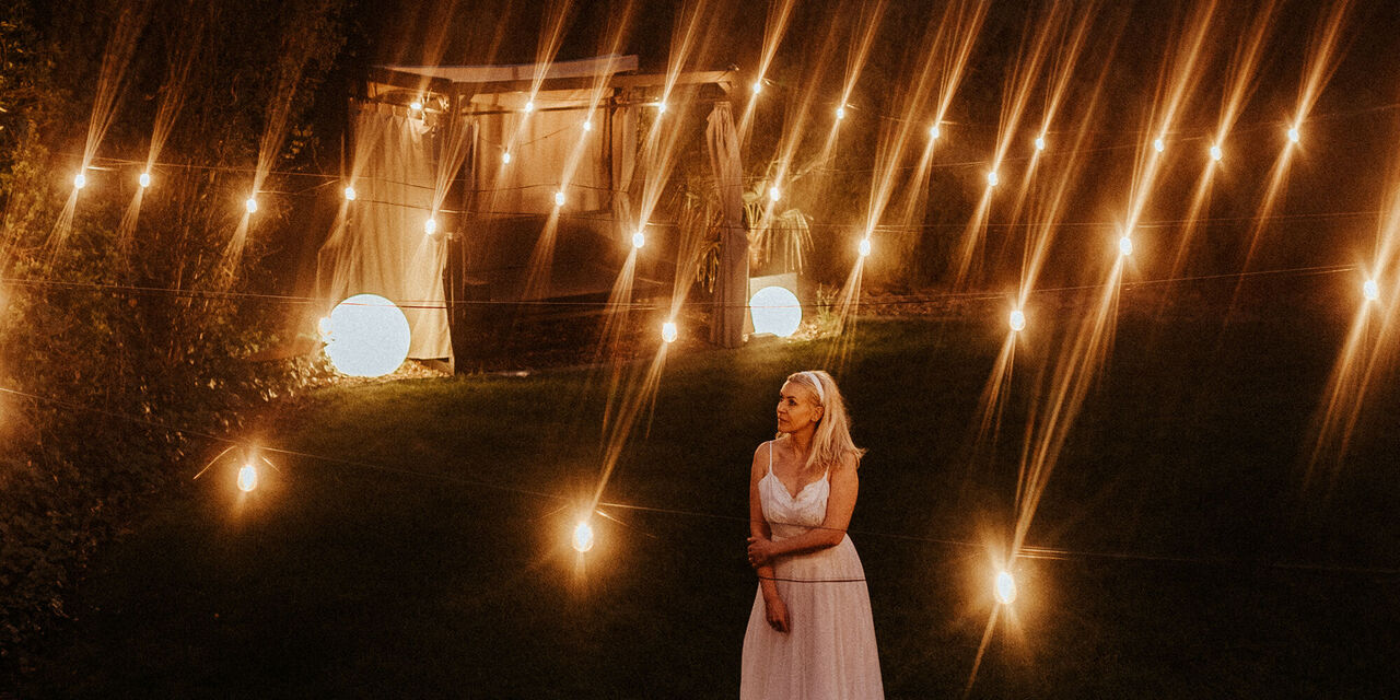 Žena u bijeloj haljini stoji pod noćnim nebom. Okružena je rasvjetom s osvijetljenim paviljonom u pozadini.
