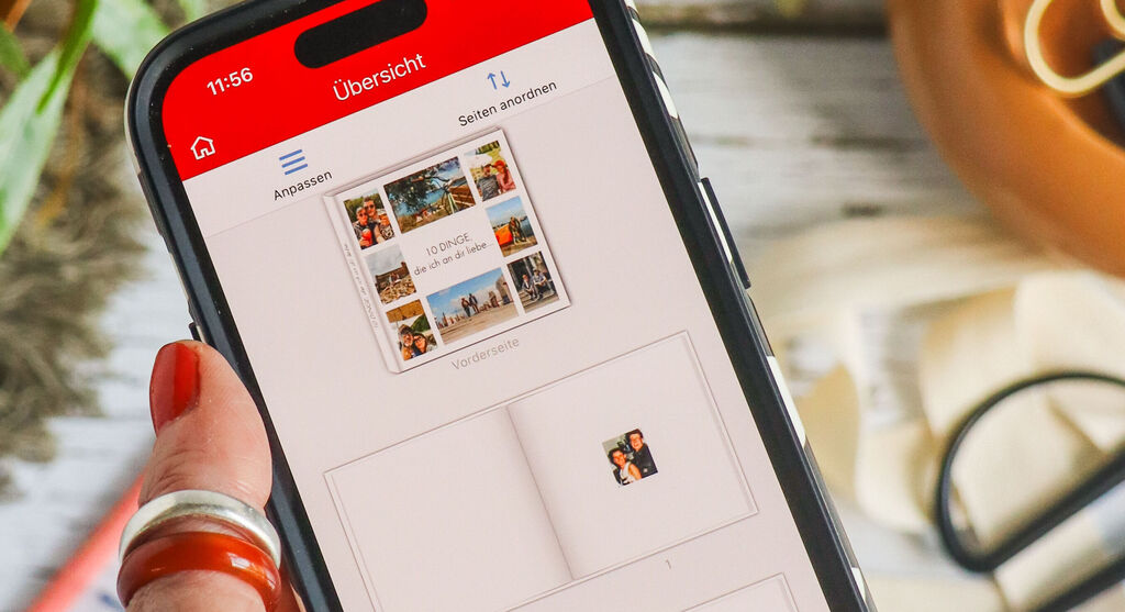 U prvom planu fotografije, ženska ruka s crvenim lakom za nokte drži pametni telefon. Na zaslonu je otvoreno sučelje za uređivanje CEWE mobilne aplikacije. Fotoalbum prikazan ranije vidljiv je u zamućenoj pozadini, leži na stolu. Zamućeni su i zelena biljka i pribor za ručni rad poput šarenih rola ljepljive trake, olovaka, škara i vrpce.